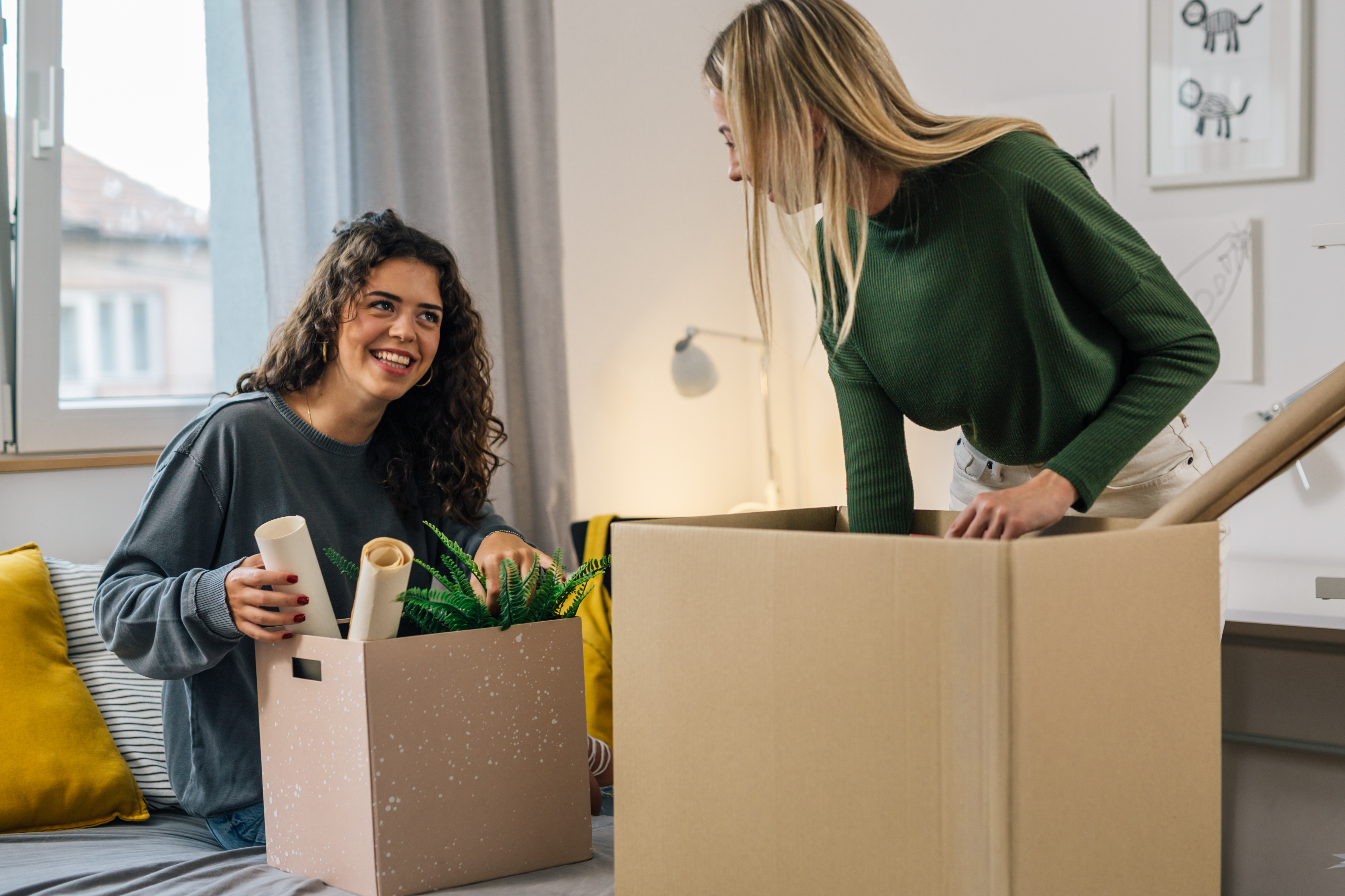 To kvinder pakker flyttekasser i en stue, én sidder og smiler, den anden står op og lager.