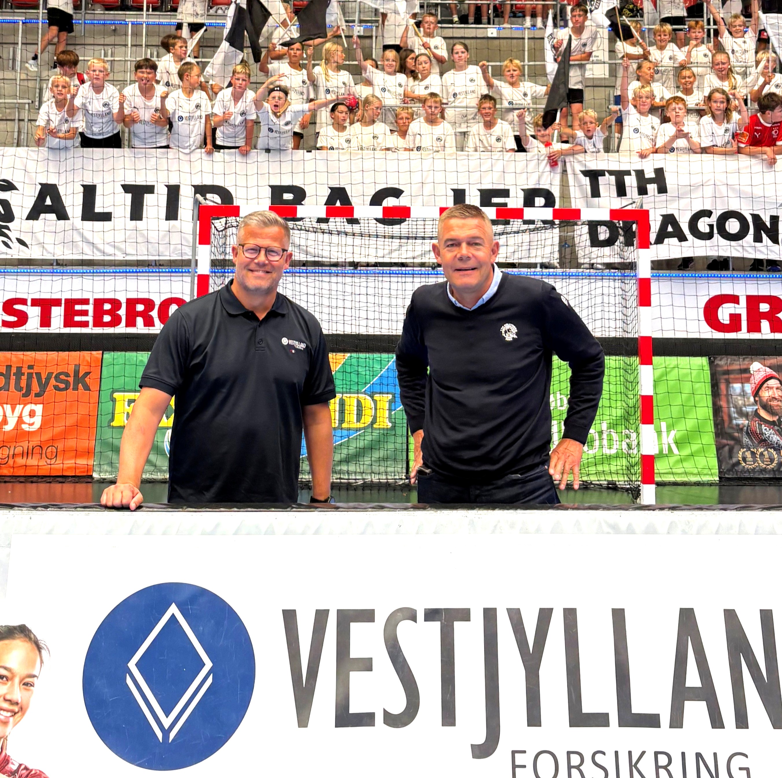 Fire mænd står udendørs med bolde, t-shirts og jakker med sponsorlogoer, foran en blomsterbede og biler.